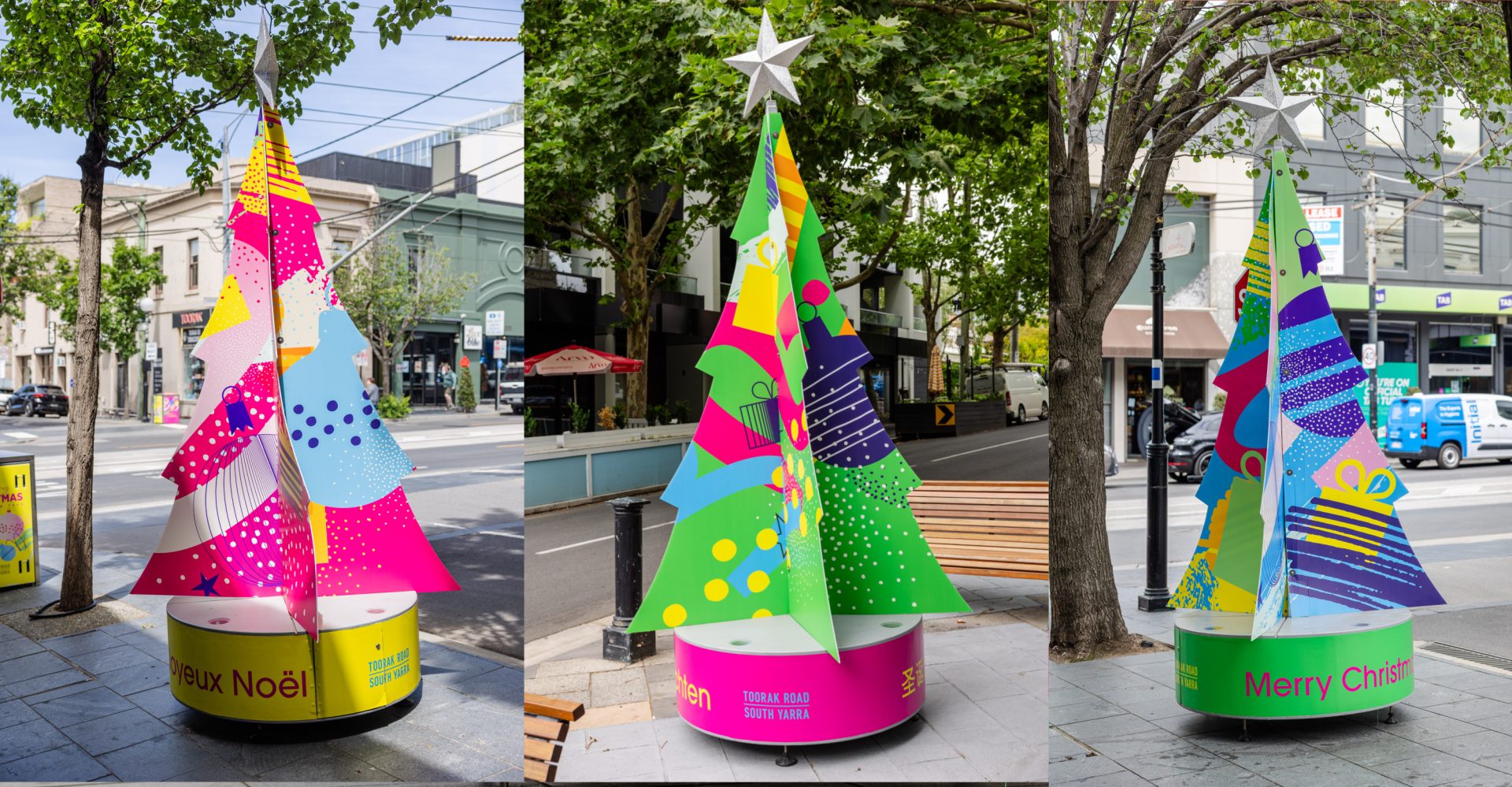 Three brightly coloured Christmas trees installed along Toorak Road South Yarra. Each tree features bold graphic patterns in pink, green, yellow and blue, topped with a silver star. The trees sit on circular bases with festive greetings in different languages, surrounded by street trees, footpaths, benches and shopfronts, reflecting a lively inner-city streetscape.