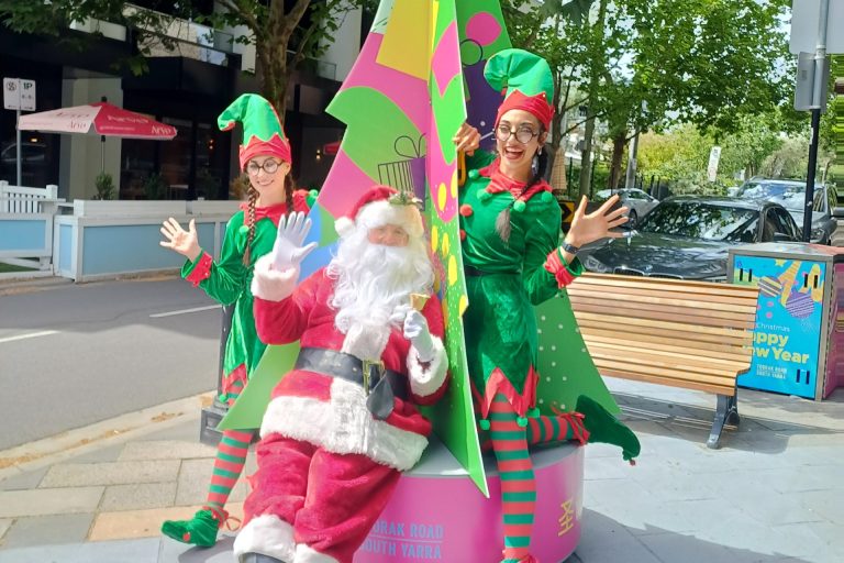 Santa sits at the base of a bright, colourful Christmas tree installation on Toorak Road, waving to the camera. Two cheerful elves stand beside him, smiling and posing with raised hands. Summer greenery, parked cars and a festive TRSY street decoration can be seen in the background.